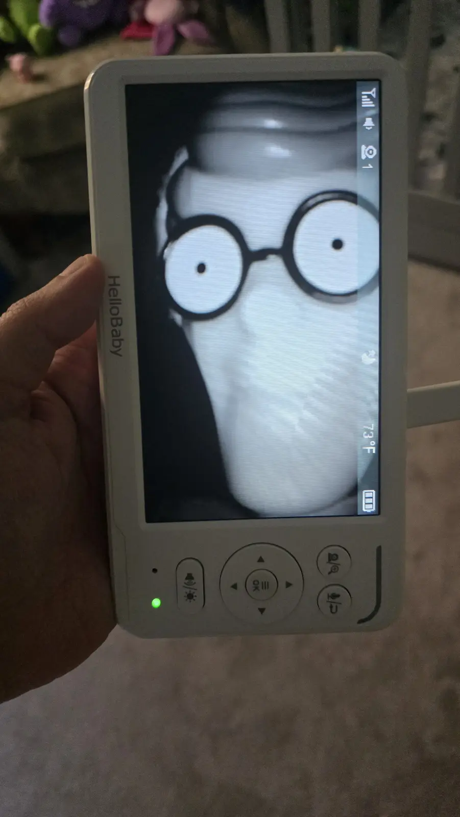 A hand holds a white HelloBaby baby monitor displaying a black-and-white image of a cat with cartoonishly large round…
