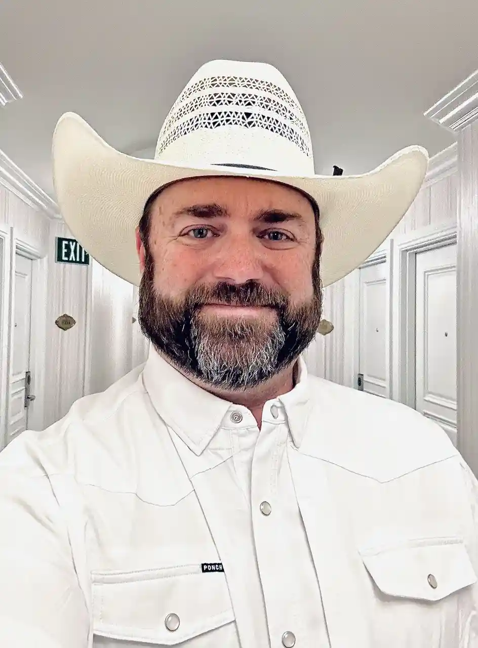 A man with a beard and blue eyes wears a large white cowboy hat and a white button-up shirt with a 'PONCH' logo, standing in…