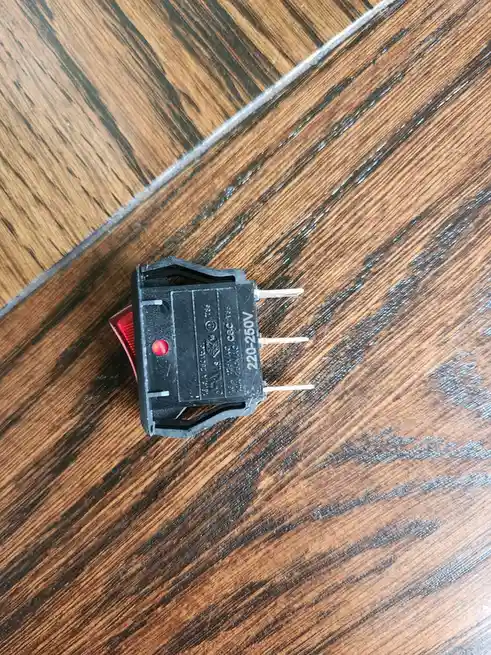 A close-up photo shows a small black electrical toggle switch with a red lever resting on a dark wood-grain surface. The…