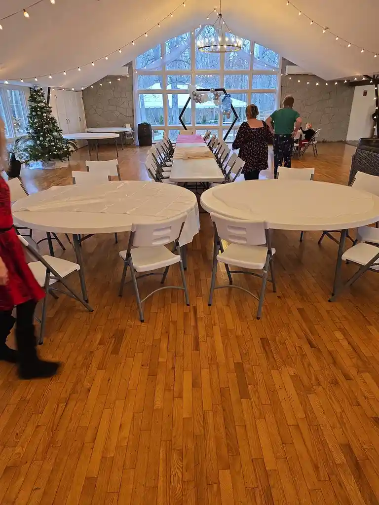 A wide shot of a festively decorated event hall with wooden floors, white folding chairs, and round tables covered in white…
