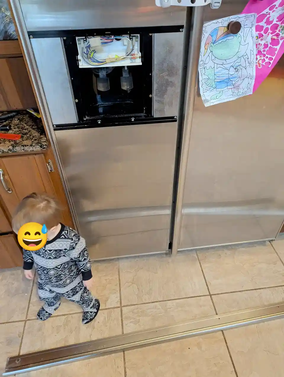 A toddler in patterned pajamas stands in a kitchen, looking up at a stainless steel refrigerator with its water and ice…