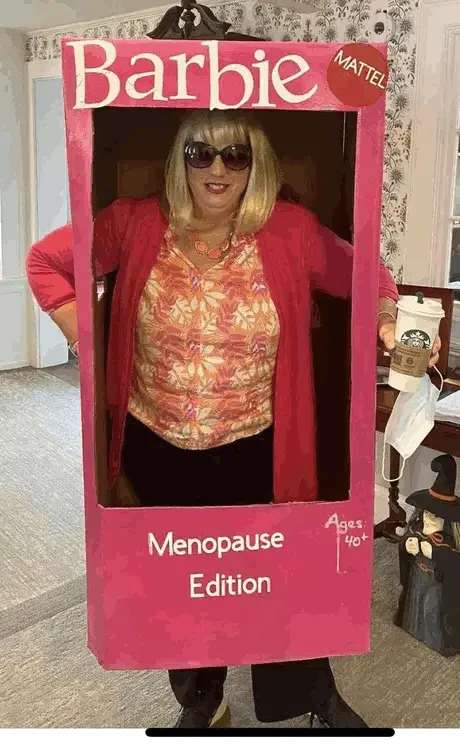 A woman wearing a pink cardboard cutout costume resembling a Barbie doll box, complete with 'Barbie' and 'MATTEL' branding…