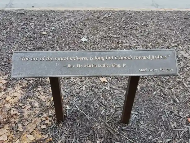 A close-up photo of a dark metal plaque set in wood chip mulch, displaying the quote 'The arc of the moral universe is long…