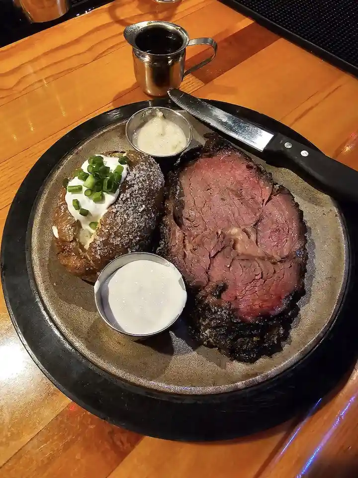 a close-up photo of a plated meal featuring sliced prime rib roast with a charred exterior and pink interior, a baked potato…