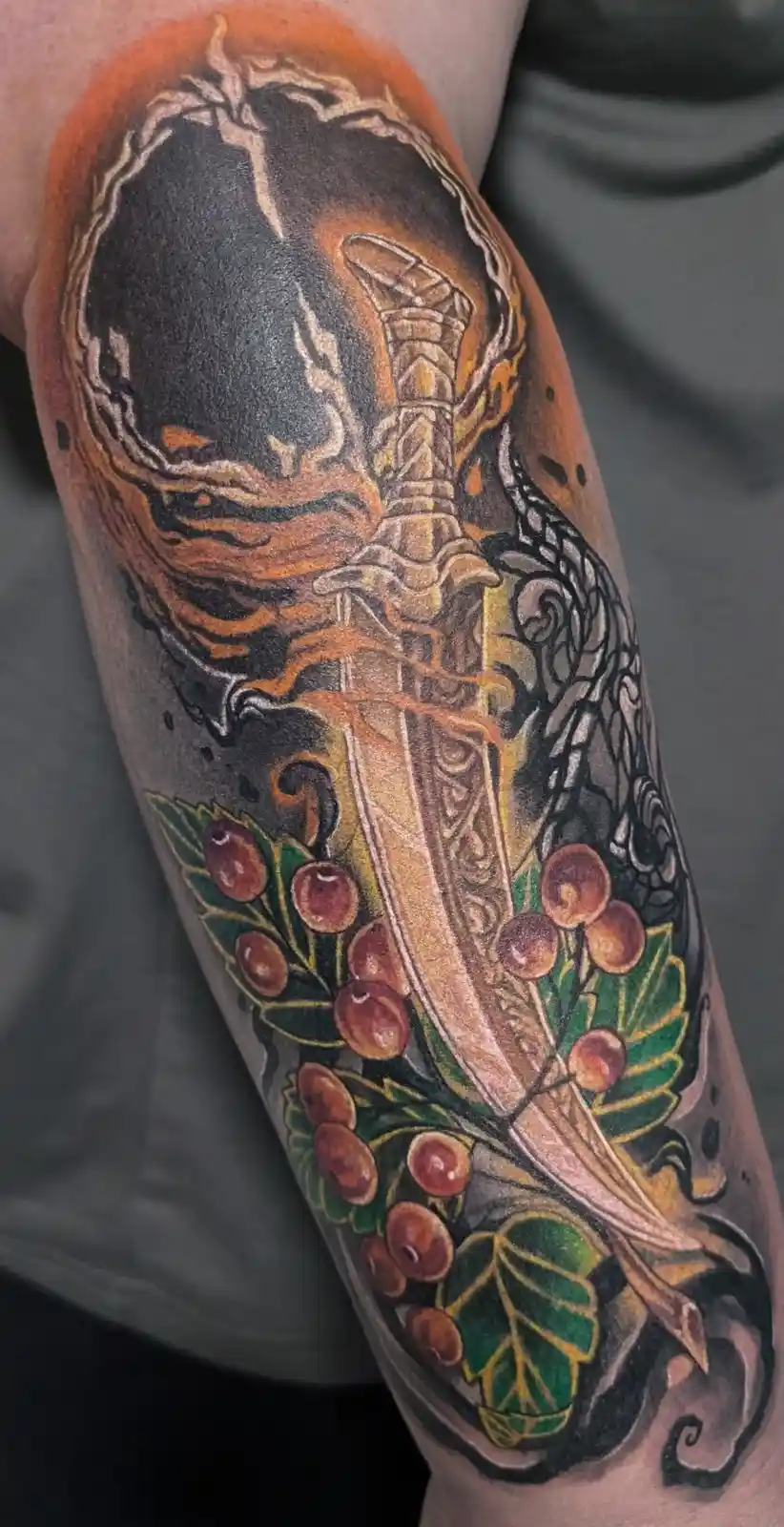 A detailed tattoo on a person's forearm depicts a sword embedded in a stone, surrounded by swirling flames and ornate…