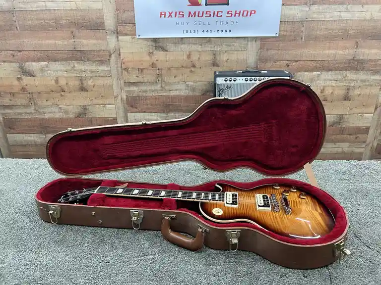 An electric guitar with a sunburst finish sits inside an open, brown hardshell case lined with red velvet, placed on a gray…