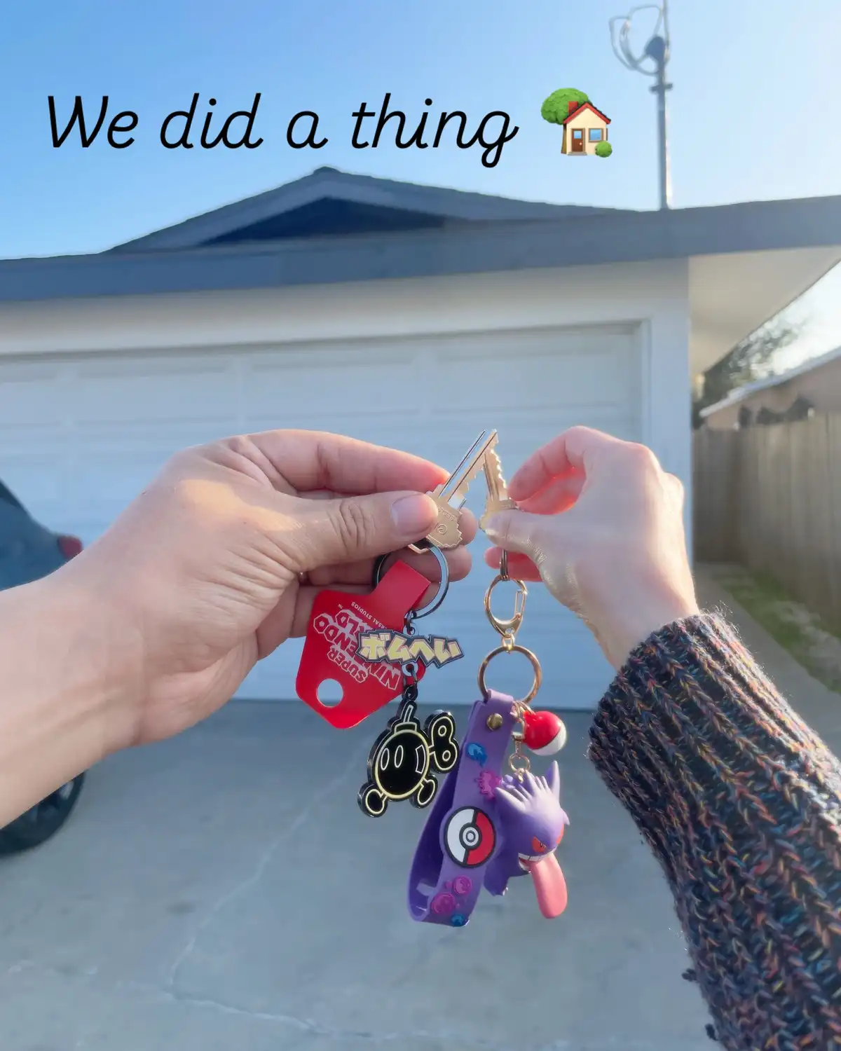 Two hands hold up house keys with colorful keychains featuring Nintendo and Pokémon characters in front of a garage. The…