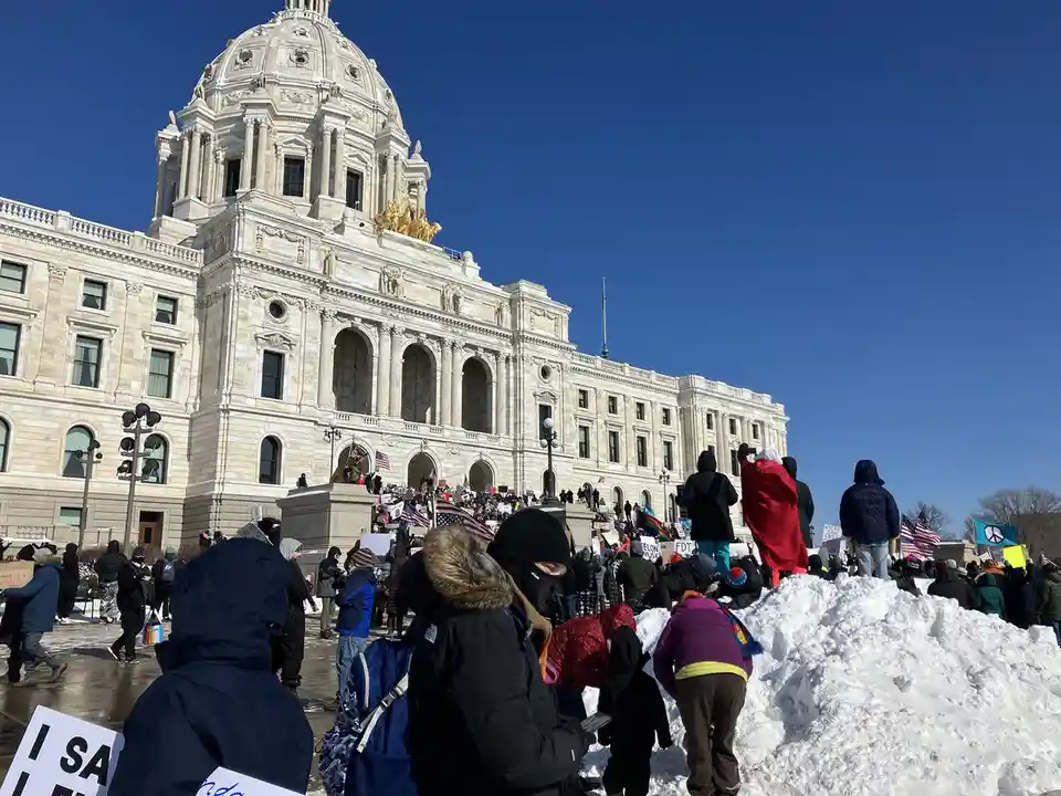 A large crowd of protesters in winter coats and hats gathers outside a grand, snow-covered state capitol building under a…