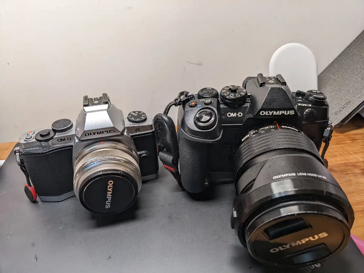 Two Olympus OM-D cameras, one silver and one black, sit side by side on a dark surface. The silver camera on the left is…