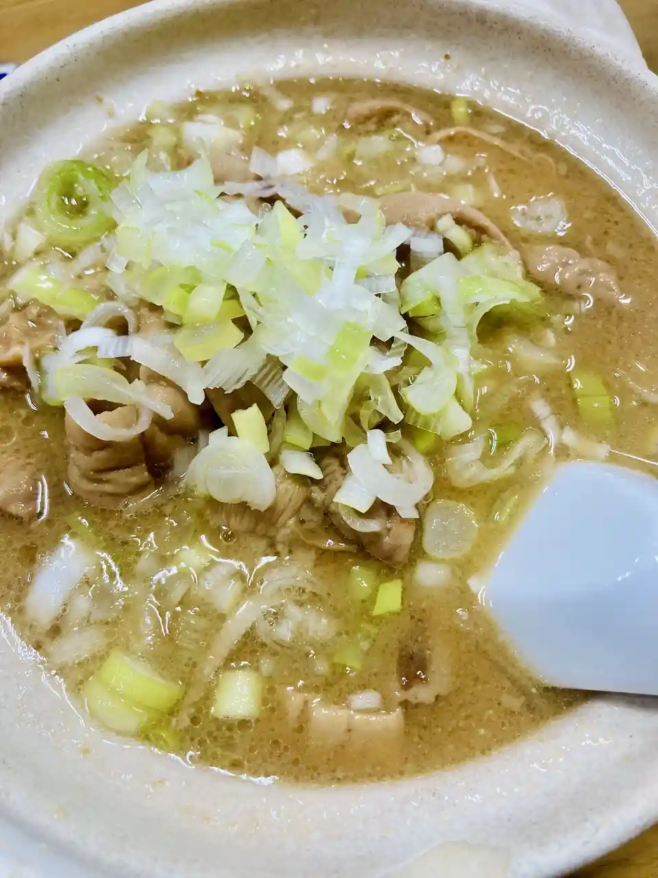 a close-up photo of a steaming bowl of ramen with a light brown broth, topped with a generous amount of finely chopped green…