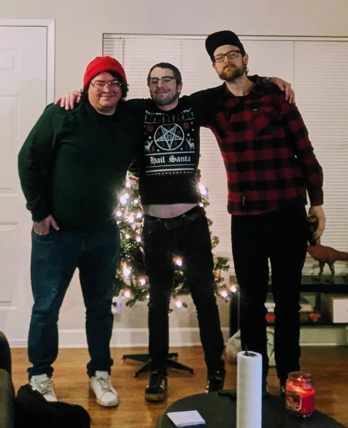 Three men pose together indoors in front of a lit Christmas tree; the center man wears a black sweater with a pentagram and…