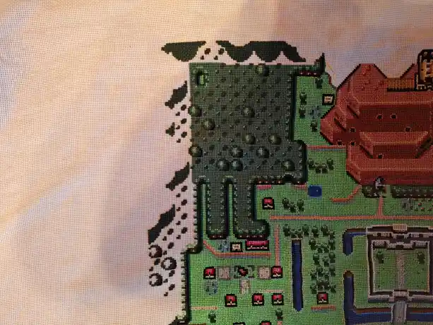A close-up of a cross-stitch artwork depicting a pixelated, top-down view of a Pokémon game map, featuring green grass…