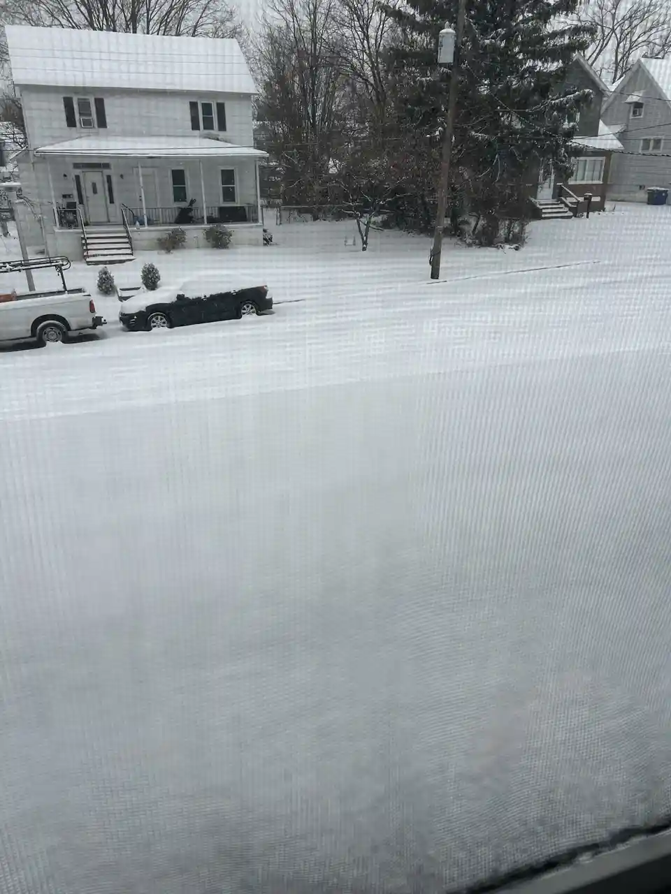A snowy residential street viewed through a window screen, with snow-covered cars and houses in the background. The overlaid…