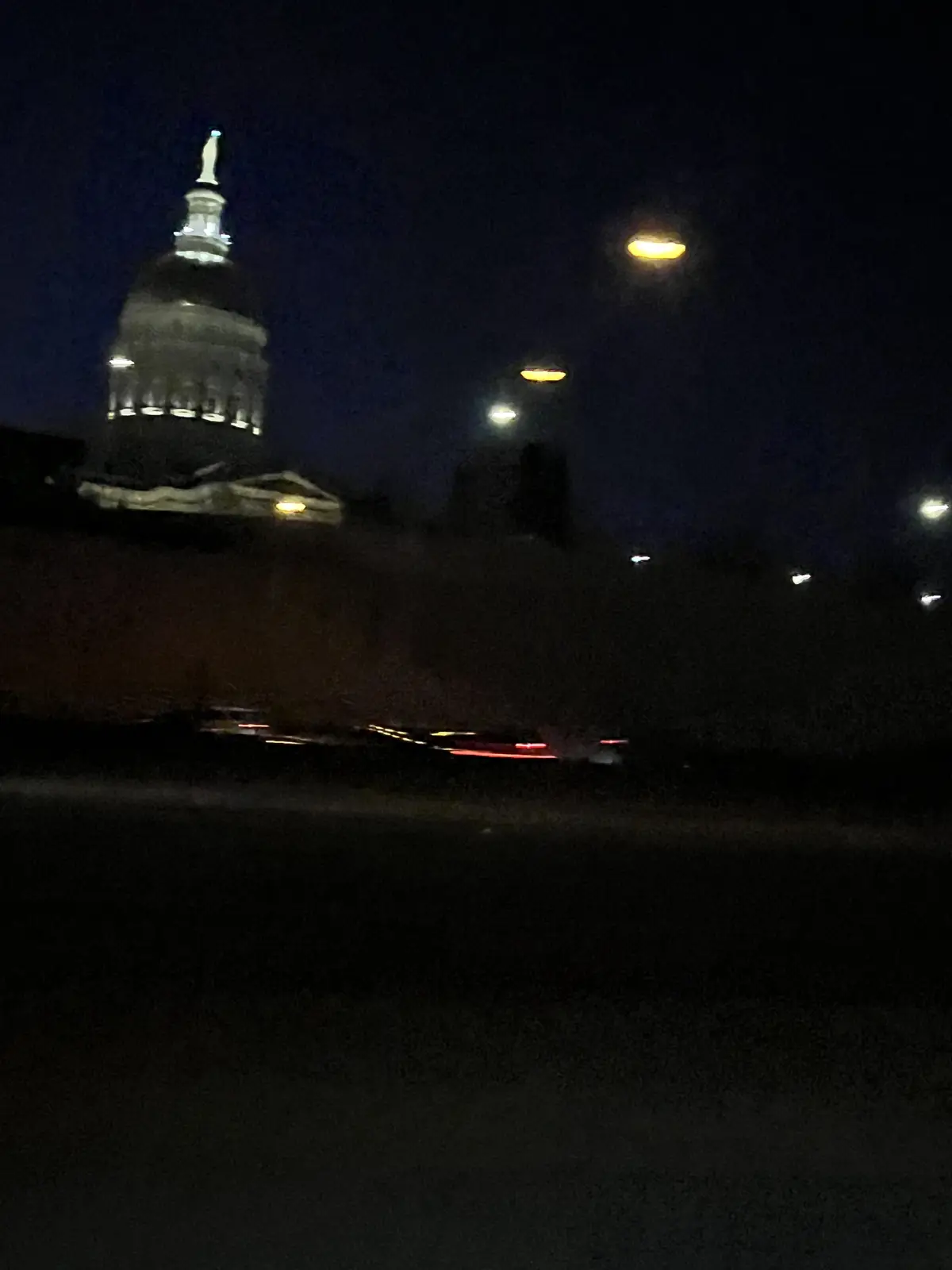 A blurry nighttime photo shows a domed capitol building illuminated against a dark sky, with streetlights and streaks of car…