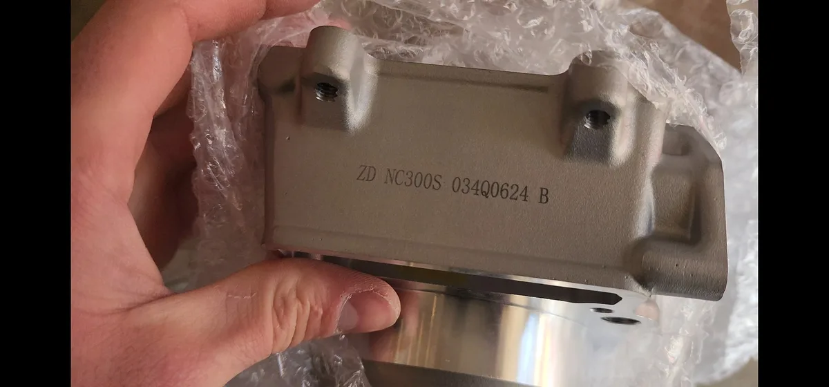 A hand holds a metallic, rectangular mechanical part wrapped in bubble wrap, showing engraved text 'ZD NC300S 034Q0624 B' on…