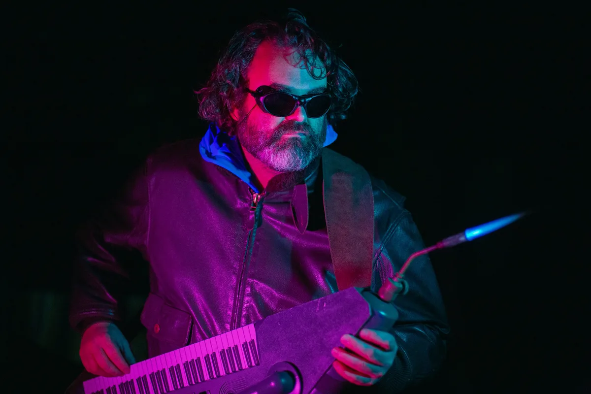 A man with a beard and sunglasses, wearing a leather jacket, plays a keyboard under dramatic red and blue lighting. A lit…
