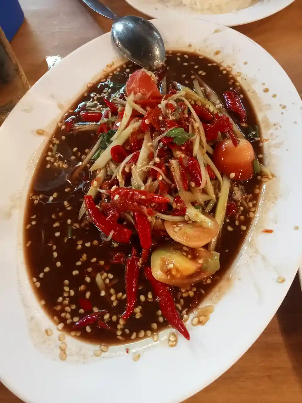 A close-up photo of a white plate holding a Thai-style spicy salad, likely papaya salad, with visible red chilies, sliced…