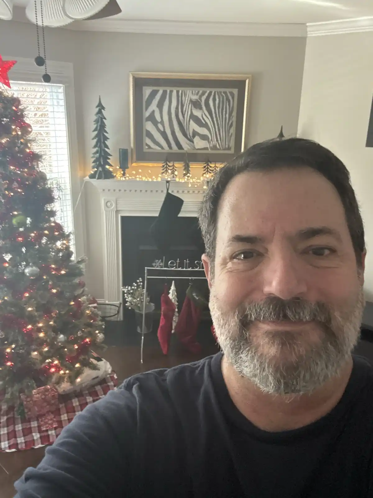 A man with a gray beard smiles at the camera in a selfie taken in a living room decorated for Christmas. To his left is a…