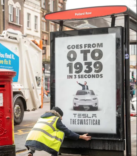 A person in a high-visibility vest is seen from behind, kneeling and touching a bus stop advertisement. The ad features a…