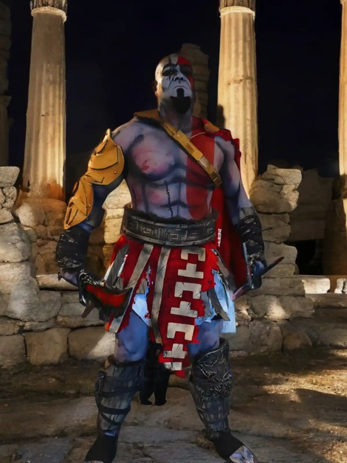 A person in a detailed Kratos cosplay from God of War stands in front of ancient stone columns at night, holding two blades…