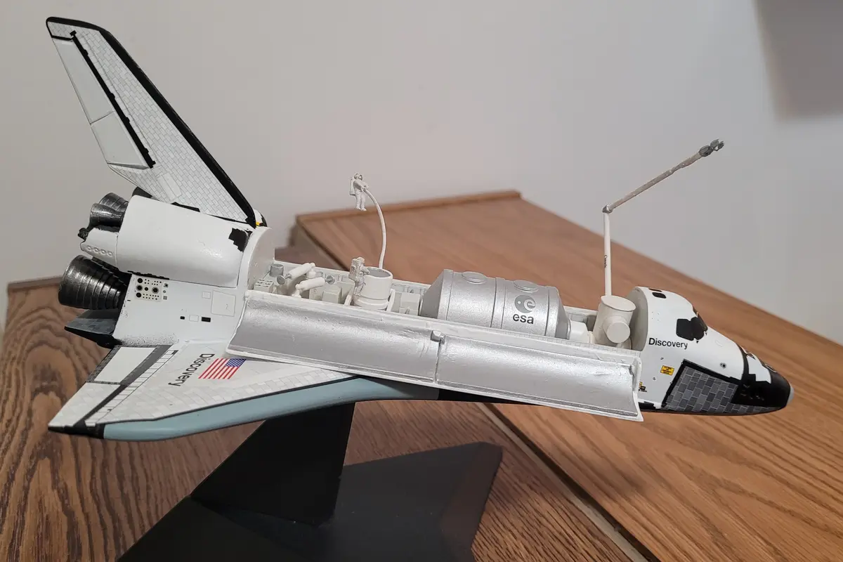 A detailed model of the Space Shuttle Discovery is shown with its payload bay open, revealing internal components and a…