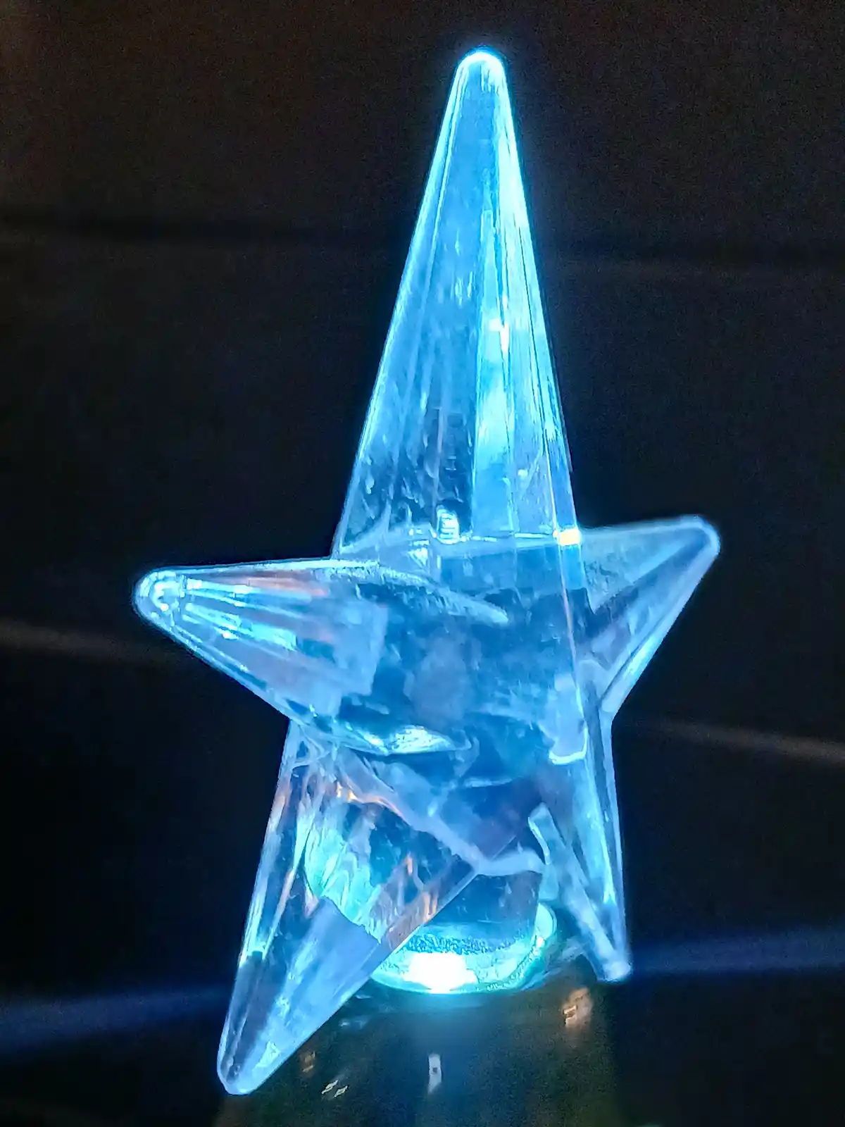 A glowing blue, translucent plastic star-shaped decoration with a pointed top, illuminated from within against a dark…