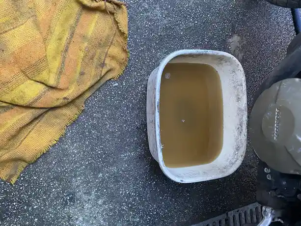 A top-down photo shows a white bucket filled with brownish motor oil sitting on a speckled gray floor next to a yellow and…