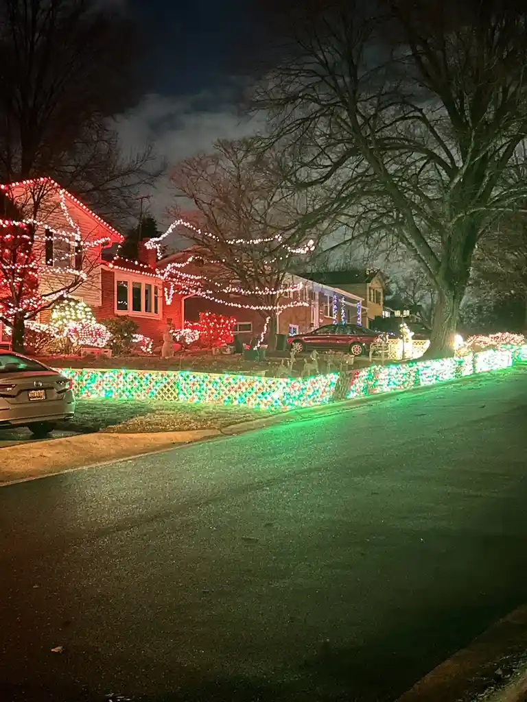 A nighttime photo of a residential street where a house is extravagantly decorated with bright red, white, and green…