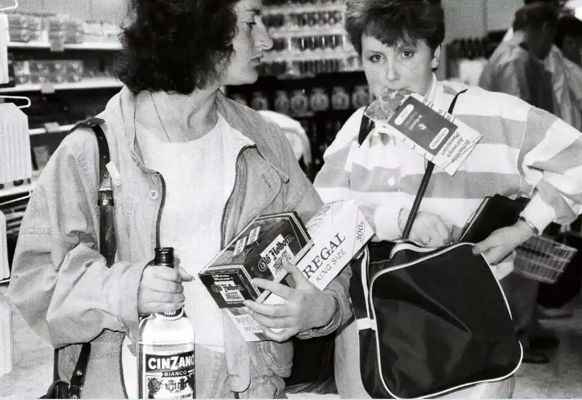 A black-and-white photo shows two women in a store aisle. The woman on the left holds a bottle of Cinzano Bianco and a pack…
