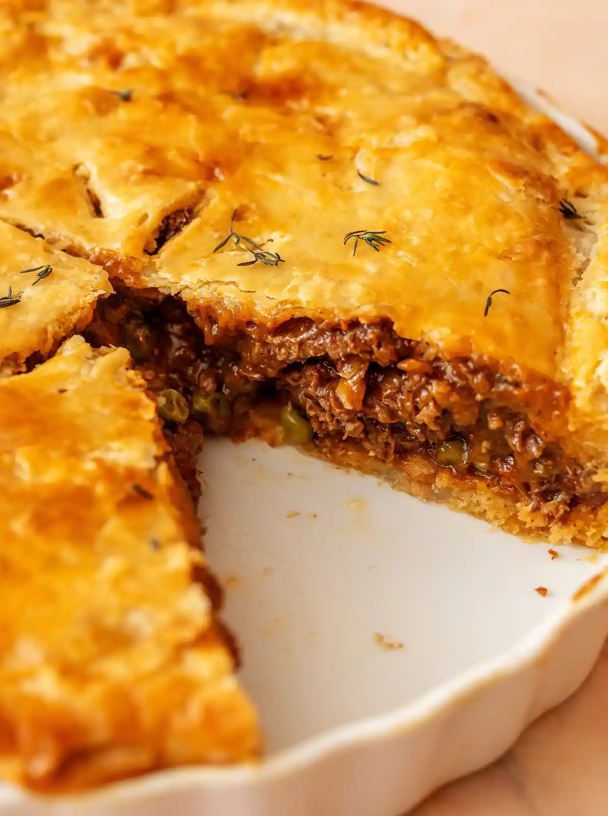 A close-up photo of a golden-brown, flaky-crust meat pie in a white ceramic dish, with a slice removed to reveal a savory…