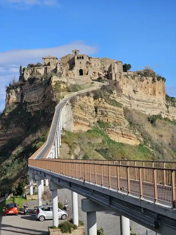 a steep, modern concrete bridge with brown railings ascends a sheer, layered cliff face to reach an ancient stone village…