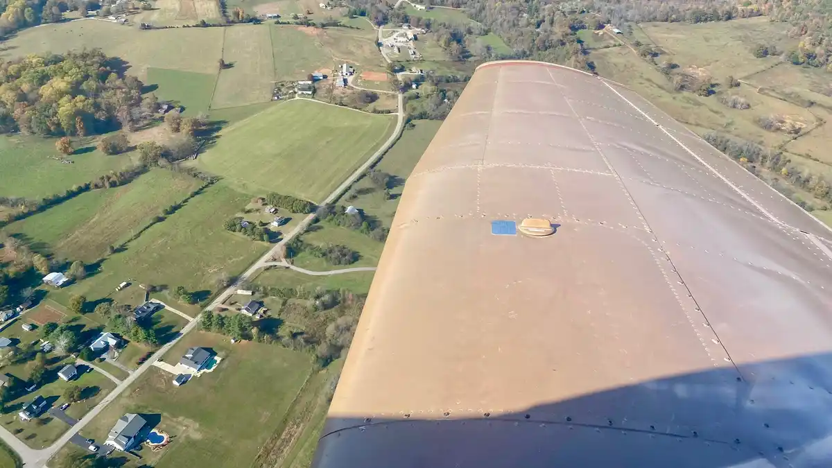 An aerial view from inside an airplane shows the wing's underside and a patchwork of green fields, roads, and houses below…