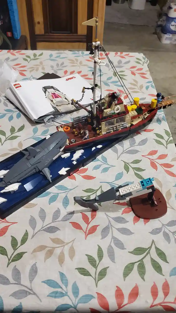 A completed LEGO Jaws movie set featuring a detailed red and beige boat with minifigures, a large grey shark model partially…