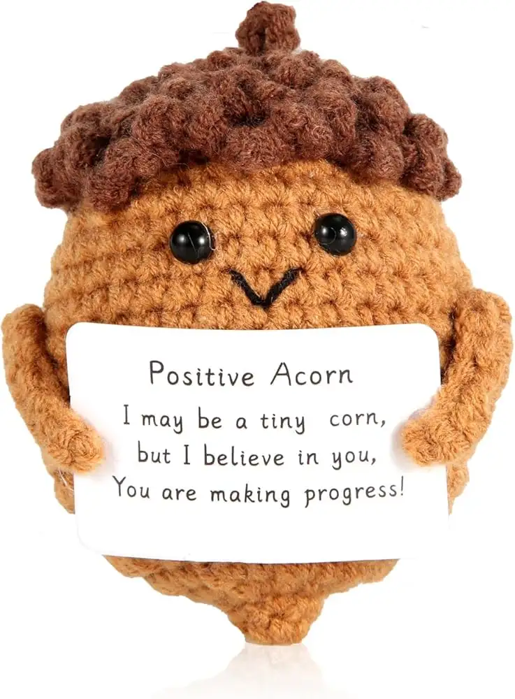 A handmade, crocheted acorn plush toy with a smiling face and brown, curly hair-like cap holds a white sign. The sign reads…