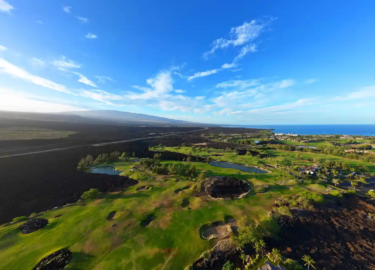 An aerial view shows a lush green golf course with sand traps and water hazards, surrounded by vast expanses of dark, barren…