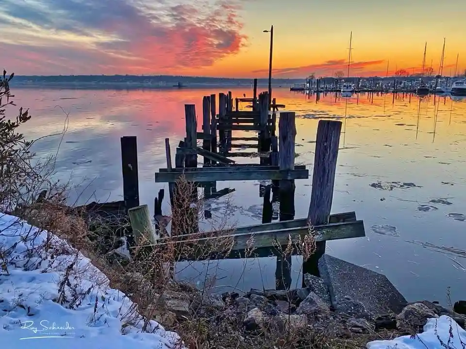 A serene winter sunset over a frozen lake with a decaying wooden pier extending into the water, snow on the shore, and…