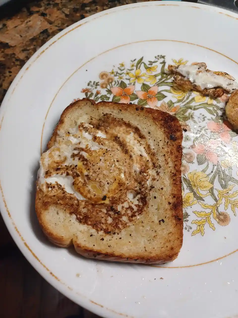 A close-up photo shows a slice of toasted bread with a fried egg cooked directly on top, seasoned with black pepper, resting…