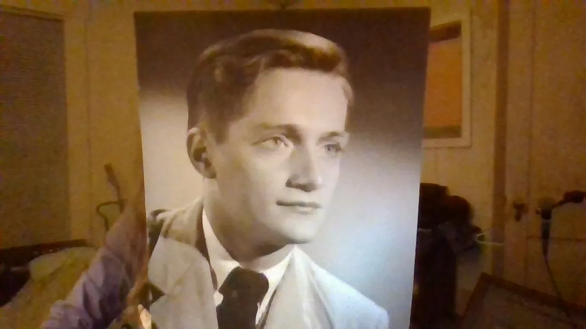 A person holds up a large, vintage black-and-white portrait of a young man in a suit and tie, looking directly at the…