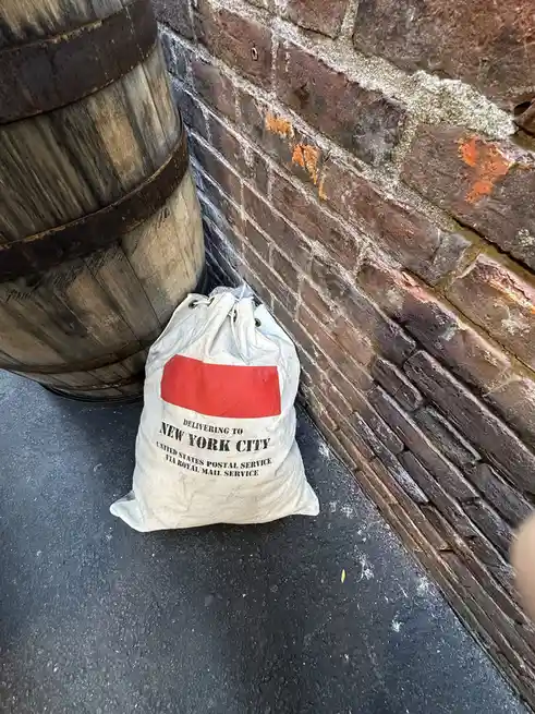 A white drawstring bag with a red stripe sits on a dark pavement against a weathered brick wall next to a wooden barrel. The…
