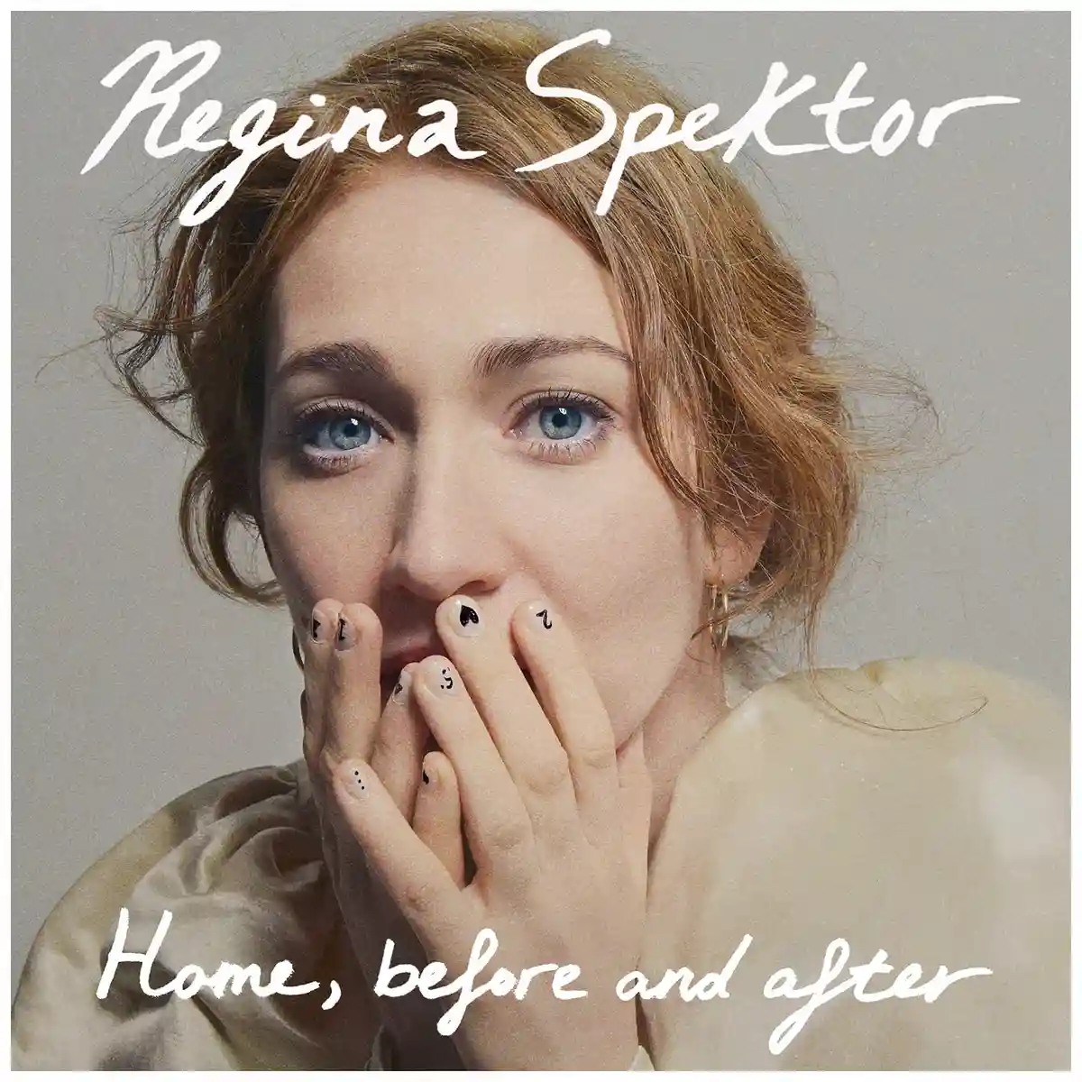 A close-up portrait of Regina Spektor with her hands covering her mouth, her fingernails decorated with small black symbols…