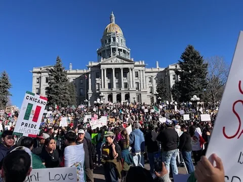A large crowd of protesters gathers in front of a grand state capitol building under a clear blue sky. Many hold signs…