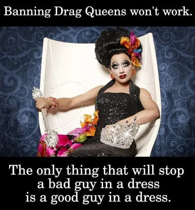 A drag queen with dramatic makeup, a large black wig adorned with colorful flowers, and a glittery black dress sits…