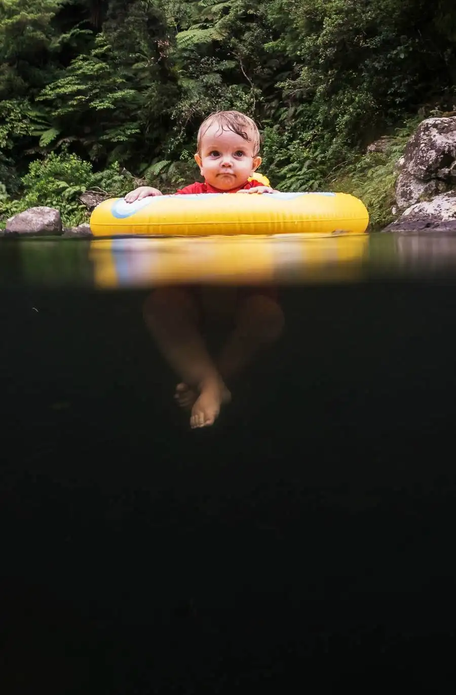 A baby in a red shirt sits in a yellow inflatable ring in a dark, calm body of water, surrounded by lush greenery and rocks…