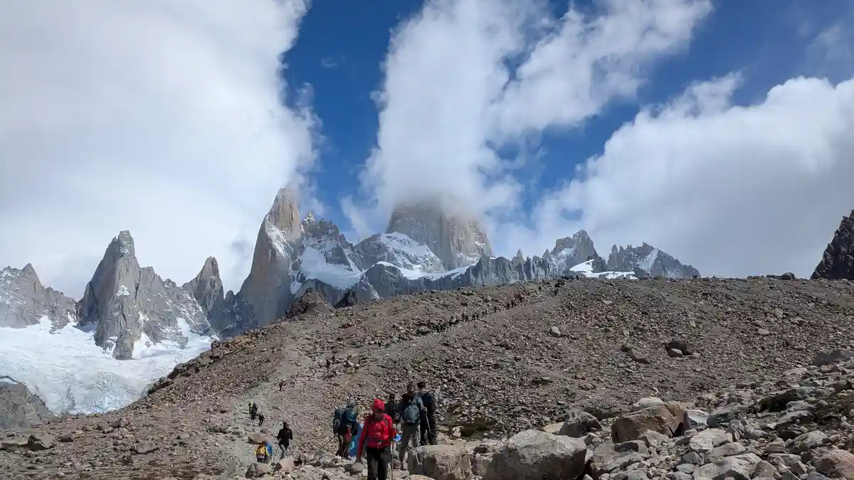 A wide shot shows numerous hikers in colorful gear trekking up a steep, rocky mountain slope under a partly cloudy sky. In…