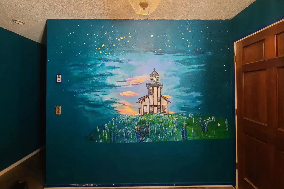 A hand-painted mural on an interior wall depicts a lighthouse on a grassy hill under a starry night sky with clouds and a…