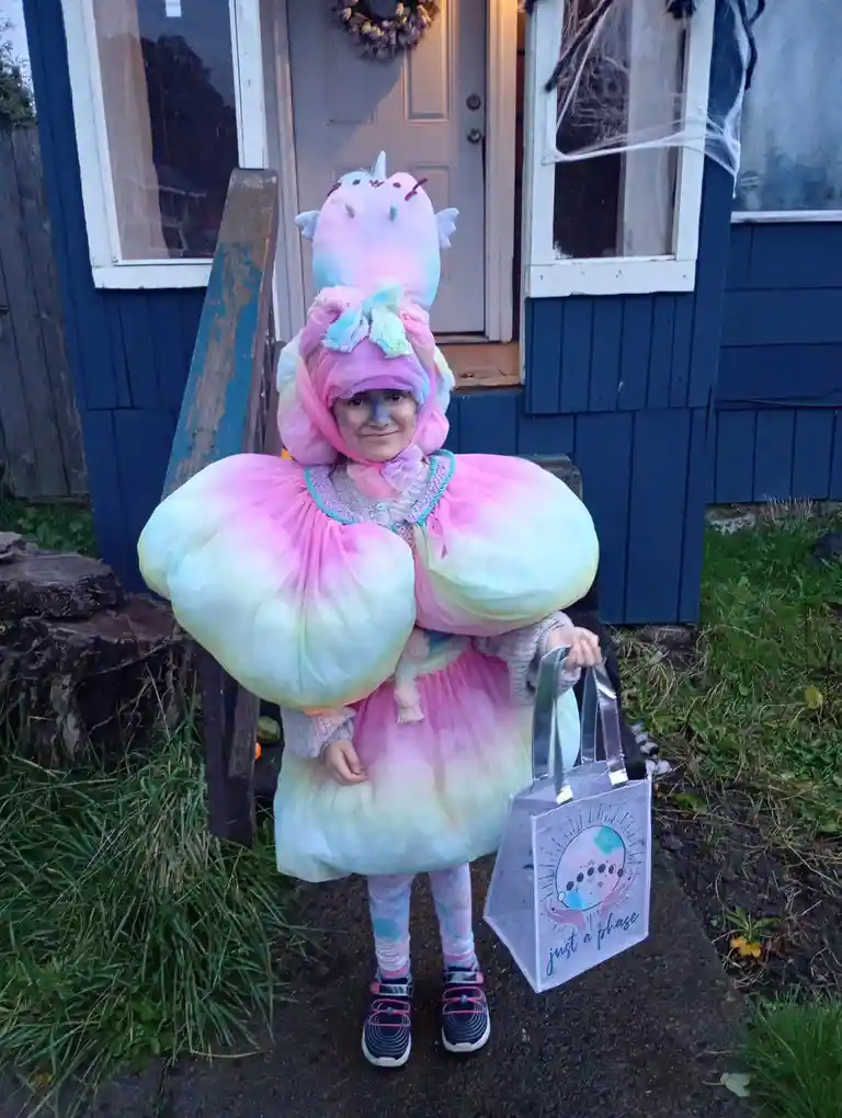 A young child in a colorful, fluffy costume resembling a Pusheen the cat unicorn stands outside a house, holding a…
