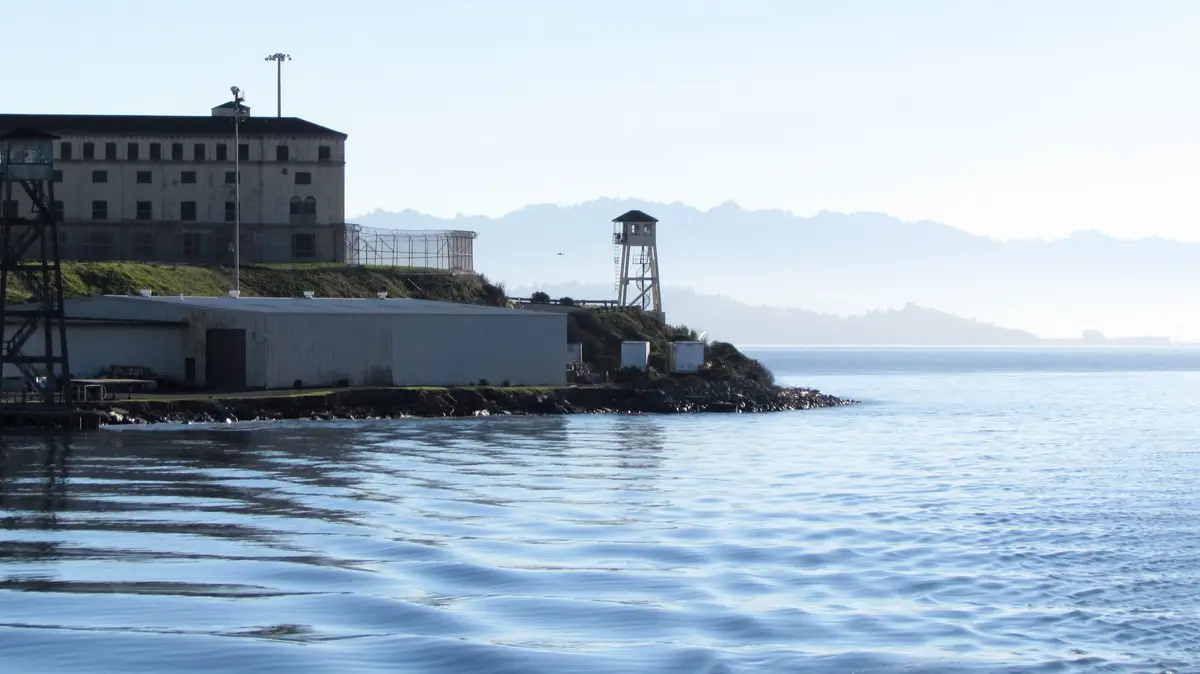 A scenic view of Alcatraz Island, showing its historic prison buildings, watchtowers, and surrounding water under a hazy…