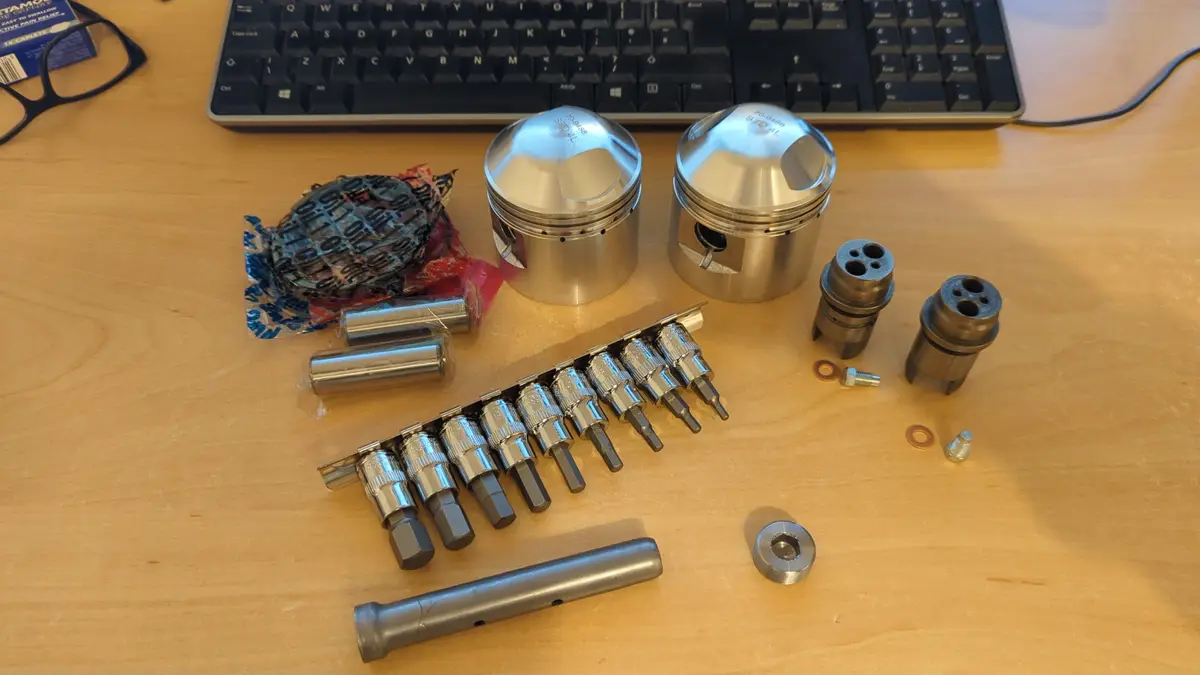 A wooden desk holds an array of shiny, complex mechanical parts including two large pistons, a set of socket wrenches, valve…