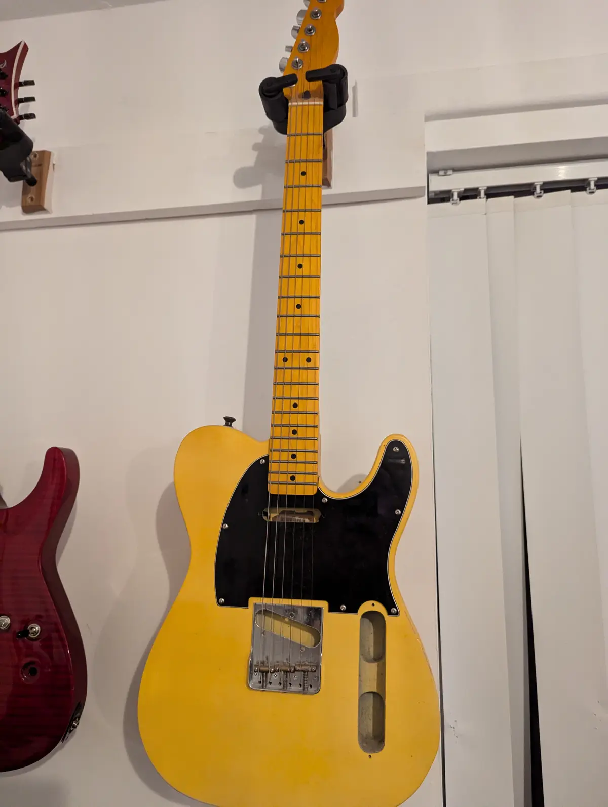 A yellow electric guitar with a black pickguard is mounted vertically on a white wall, its neck held by a black clamp. To…