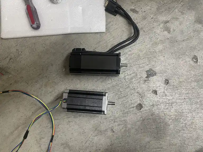 Two stepper motors lie on a concrete floor, one with a black housing and thick black cables, the other with a silver housing…